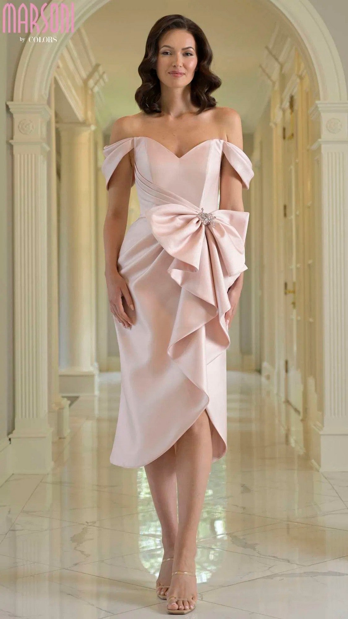 A woman stands in a hallway wearing the Marsoni MV1389 knee-length semi-formal dress in mauve with an off-the-shoulder design, oversized bow detail, and strappy heels.